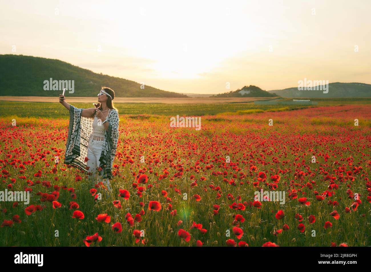 Eine Frau isst bei Sonnenuntergang in einem Mohnfeld Erdbeeren und macht Selfies auf ihrem Handy. Vorderansicht. Stockfoto