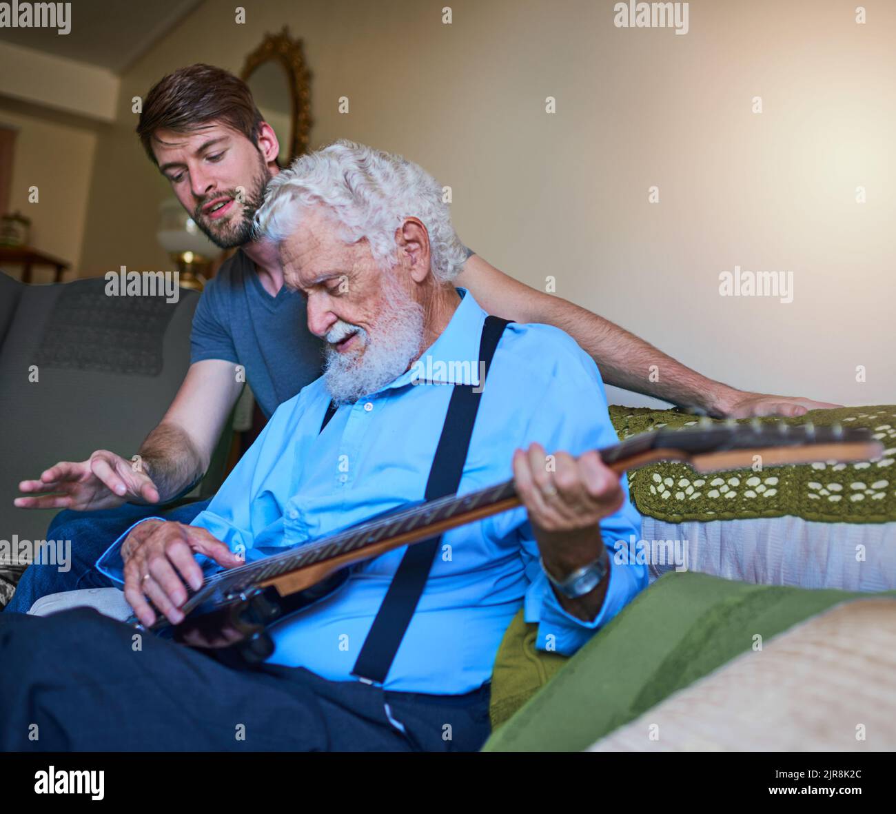 Ein glücklicher junger Mann, der seinem älteren Großvater beibringt, zu Hause auf der Couch die E-Gitarre zu spielen. Stockfoto