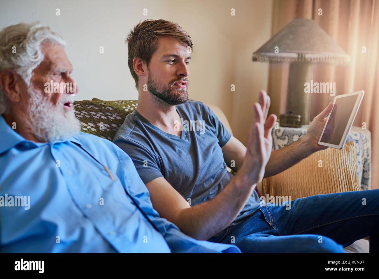 Du wirst soziale Medien lieben. Ein junger Mann zeigt seinem älteren Großvater, wie man ein Tablet benutzt, während er zu Hause auf der Couch sitzt. Stockfoto