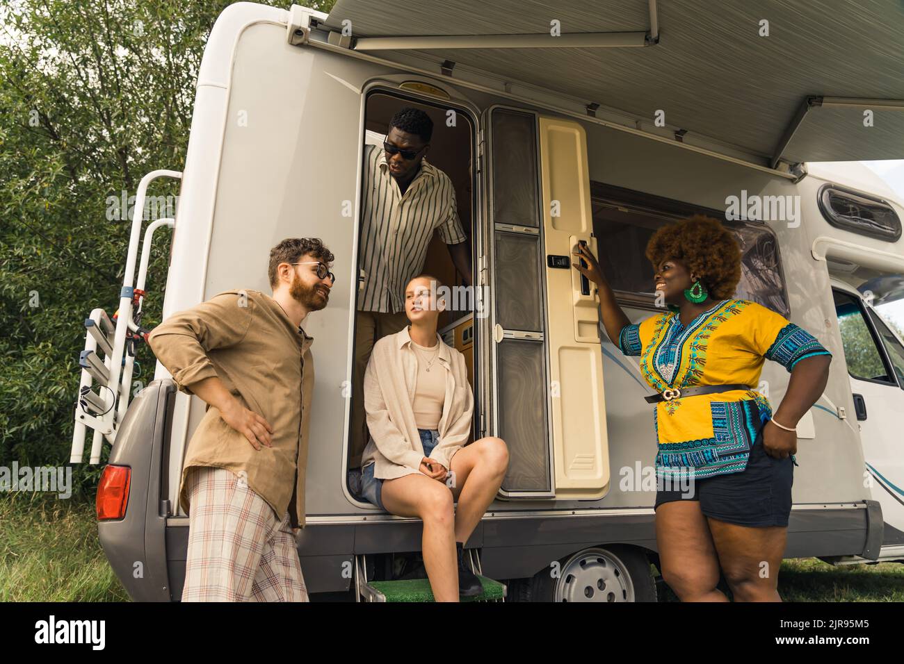 Campingurlaub im Sommer. Eine multikulturelle Gruppe von vier guten Freunden steht und redet, lacht und genießt es, Zeit zusammen in der Natur zu verbringen, während sie in ihrem komfortablen Wohnmobil leben. Hochwertige Fotos Stockfoto