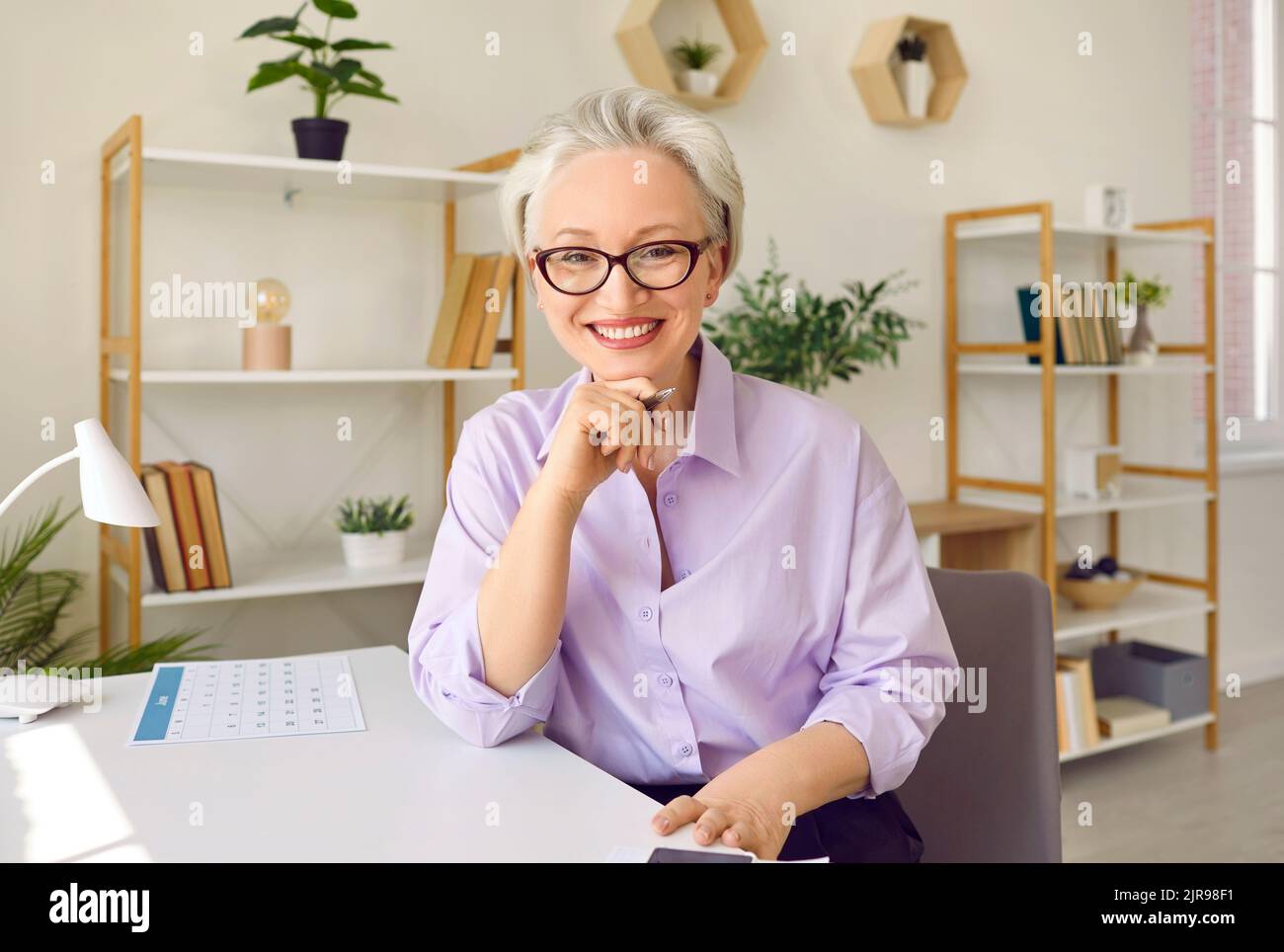 Eine ältere glückliche Frau mit grauem Haar, die lächelt und die Kamera anschaut, sitzt auf dem Schreibtisch im stilvollen Büro Stockfoto
