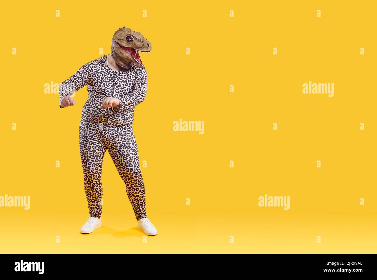 Witziger exzentrischer übergewichtiger Mann, der in einer seltsamen Dinosaurier-Gummimaske auf orangefarbenem Hintergrund tanzt. Stockfoto