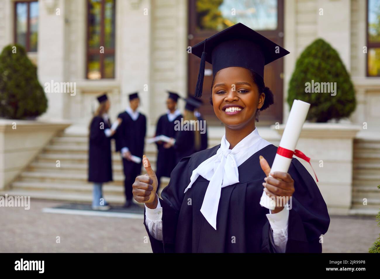 Freudige Universitätsstudentin, die ein ausgezeichnetes Zeichen zeigt, während sie mit einer Diplomrolle steht. Stockfoto