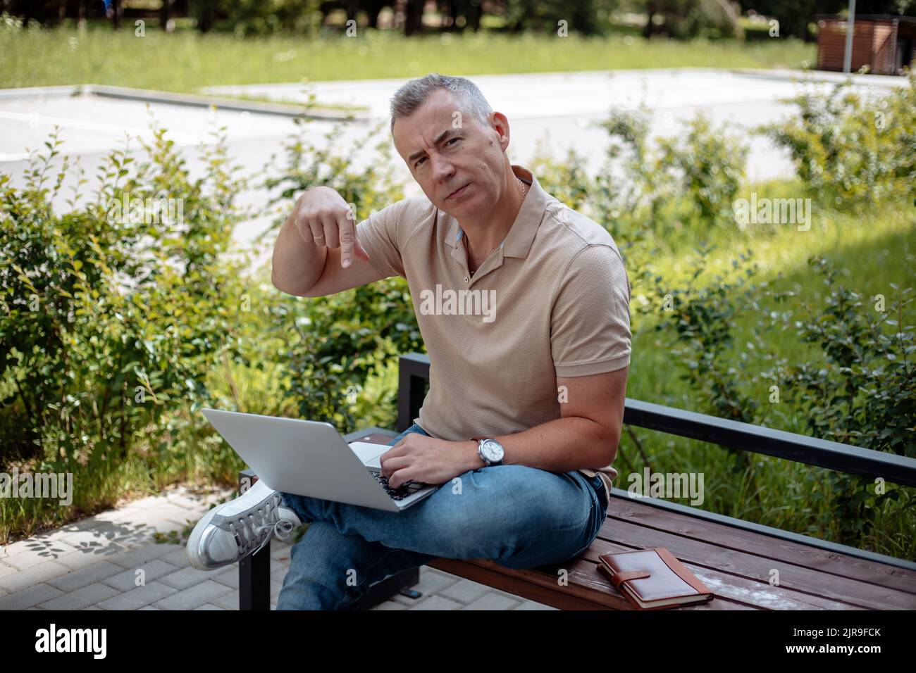 Älterer Mann, der im Sommer auf einer Holzbank mit gekreuztem Bein im Park sitzt, seinen Laptop in der Hand hält und mit dem Zeigefinger nach unten zeigt. Stockfoto