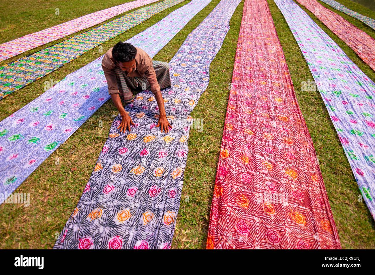 Narayanganj, Dhaka, Bangladesch. 23. August 2022. Farbenfrohe Stoffstreifen bilden ein auffälliges Display, da sie in ordentlichen Reihen auf einem Feld in Narayanganj, Bangladesch, angeordnet sind. Die langen Baumwolltücher werden lokal als ''Saree'' bezeichnet - ein traditionelles Kleidungsstück für Frauen, das unter der heißen Sonne trocknen soll und mit hellen Farben gefärbt wurde. Hier werden täglich etwa 4000 Stück Stoff zum Trocknen gelegt. Der Prozess dauert in der Regel drei Stunden, wobei jeder Satz von 200 Stück auf einmal bei Temperaturen trocknen kann, die über 36 Grad celsius erreichen können. Kredit: ZUMA Press, Inc./Alamy Live Nachrichten Stockfoto