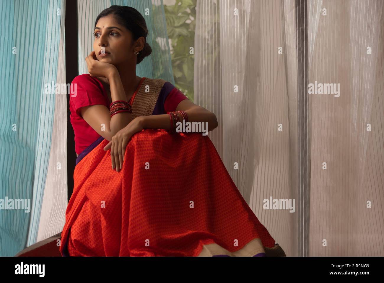 Junge Frau, die mit der Hand auf dem Kinn nach draußen blickt, während sie zu Hause neben dem Fenster sitzt Stockfoto