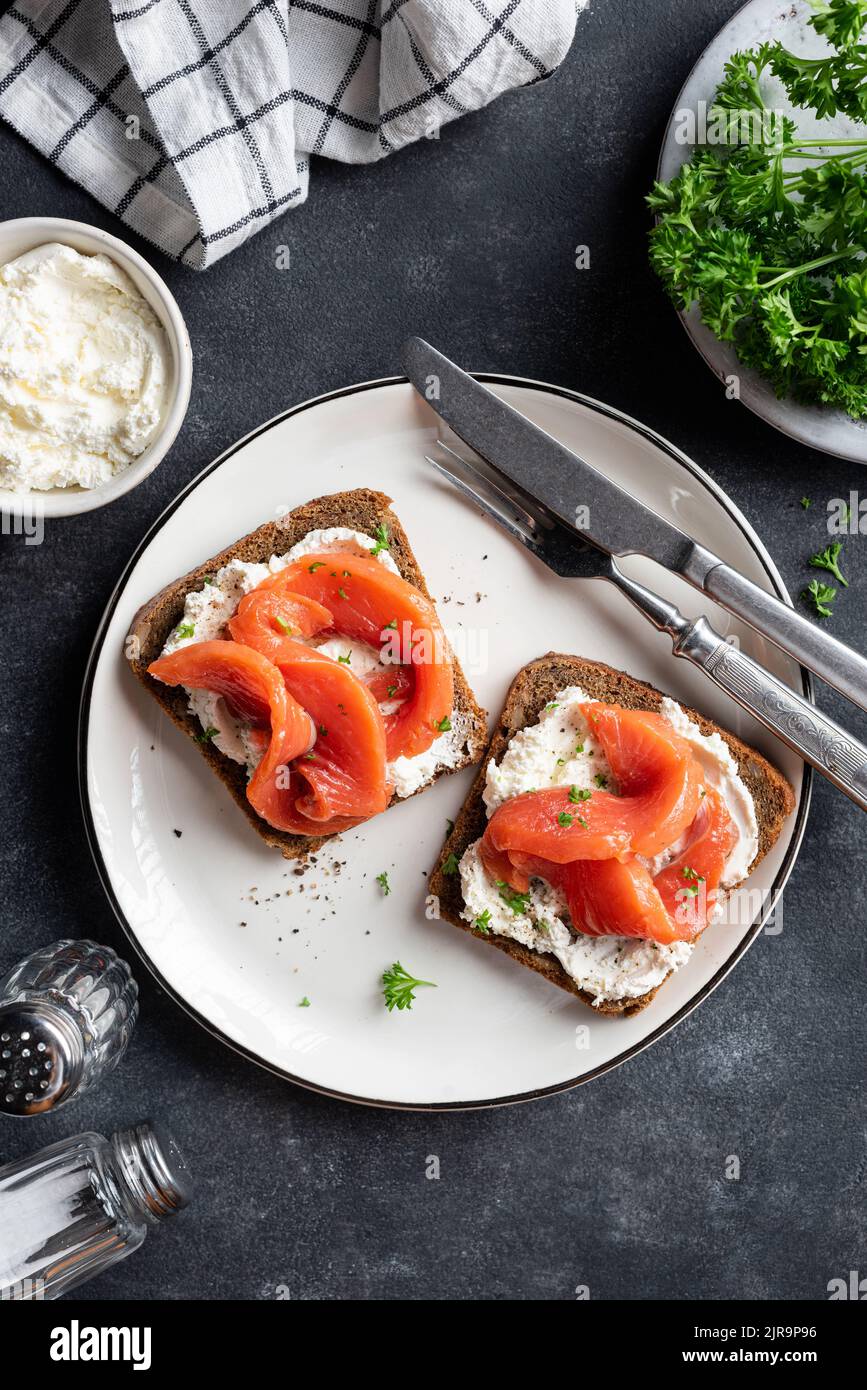 Sandwich mit geräuchertem Lachs und Frischkäse auf weißem Teller, Draufsicht, schwarzer Steinhintergrund Stockfoto