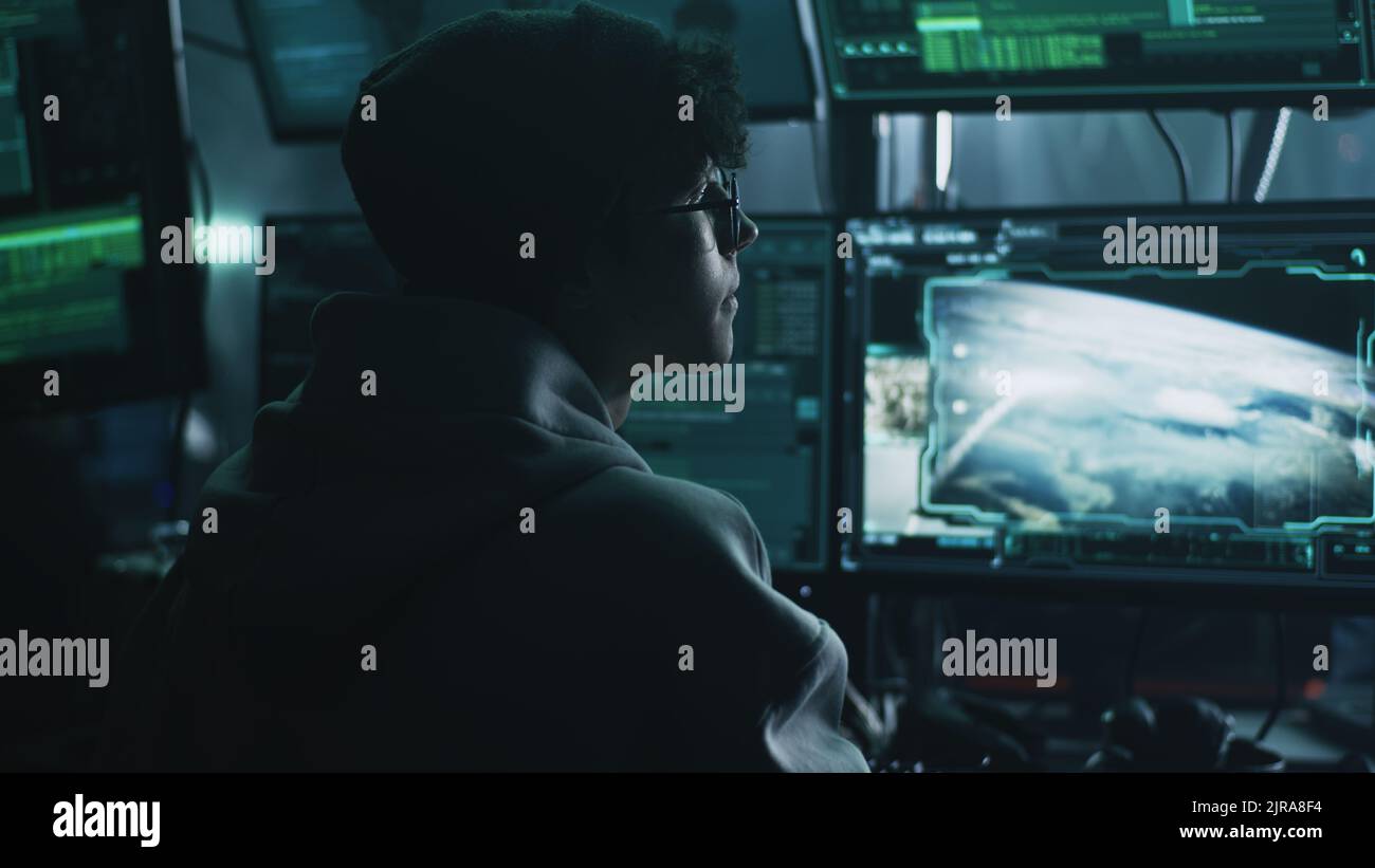 Junger, männlicher Hacker mit Hut und Brille, der Computer benutzt, um nukleare Sprengköpfe zu hacken und Krieg zu beginnen, während er am Tisch im dunklen Raum sitzt Stockfoto