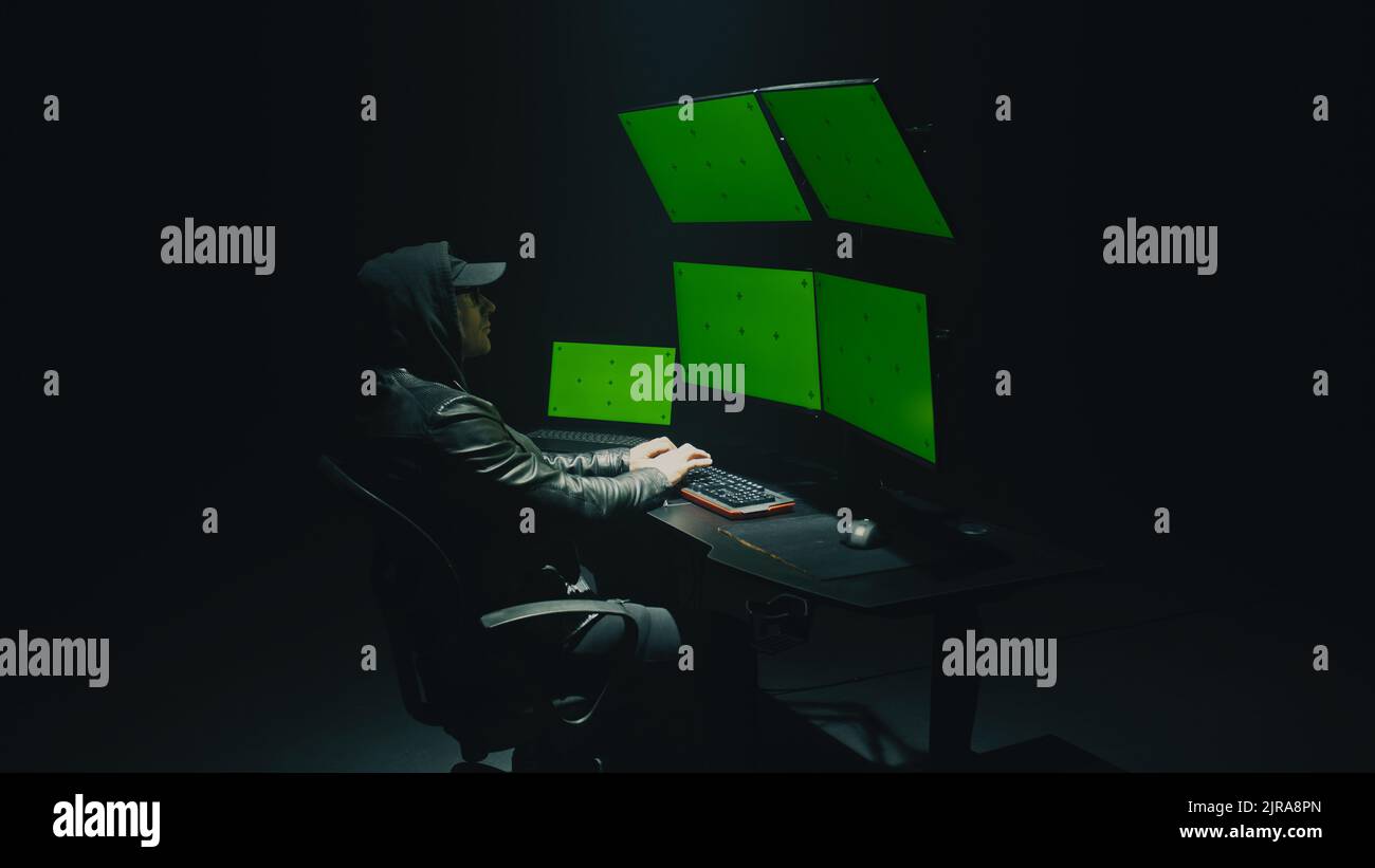 Ein männlicher Cyberkrimineller mit Brille und Kapuze hackt das Sicherheitssystem im Netzwerk und infiziert mit einem Virus in Daten auf einem Computer mit Software, in einem dunklen Raum in der Nacht Stockfoto