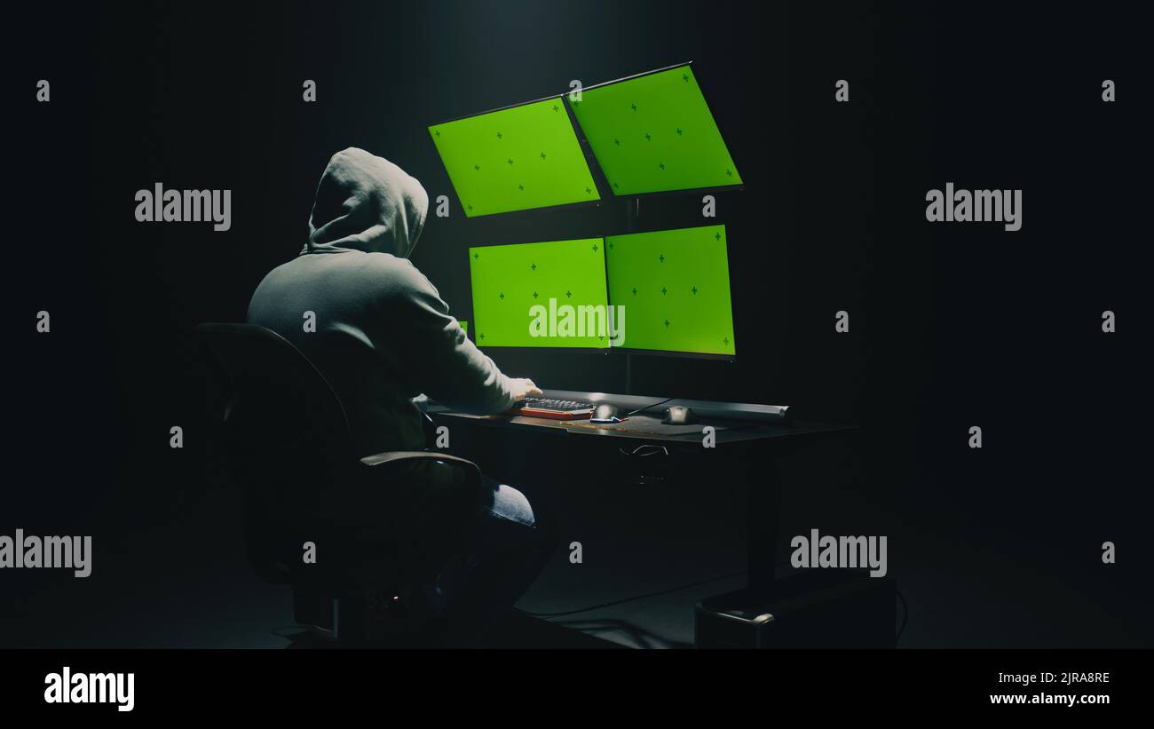 Konzept Aufnahme von anonymen Mann im Hoodie schreiben Code, um Datenbank zu hacken, während am Schreibtisch mit Computermonitoren sitzen, mit grünen Bildschirmen chromakey in dunklen Raum der Hacker-Basis Stockfoto