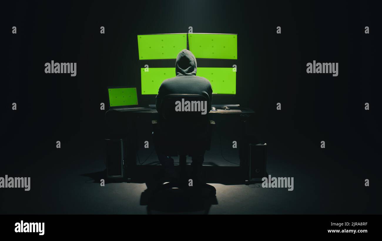 Konzept Aufnahme von anonymen Mann im Hoodie schreiben Code, um Datenbank zu hacken, während am Schreibtisch mit Computermonitoren sitzen, mit grünen Bildschirmen chromakey in dunklen Raum der Hacker-Basis Stockfoto