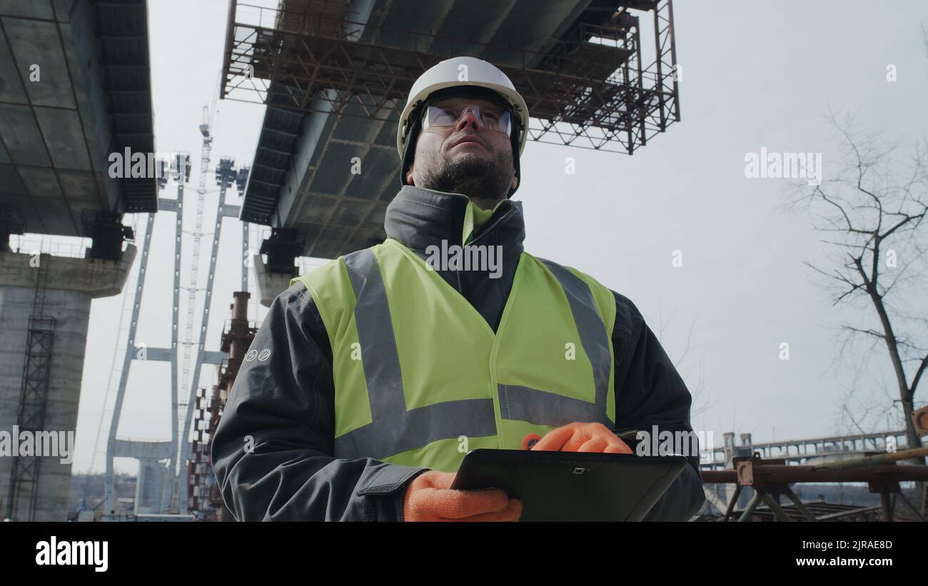 Männlicher Baumeister, der während der Bauarbeiten auf der Baustelle unter einer unfertigen Brücke steht, Daten auf dem Tablet durchstöbert Stockfoto