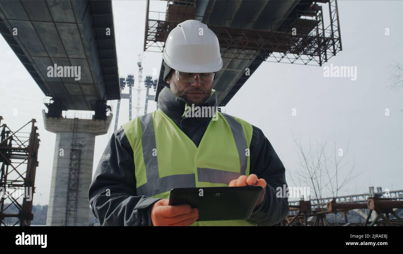 Männlicher Baumeister, der während der Bauarbeiten auf der Baustelle unter einer unfertigen Brücke steht, Daten auf dem Tablet durchstöbert Stockfoto
