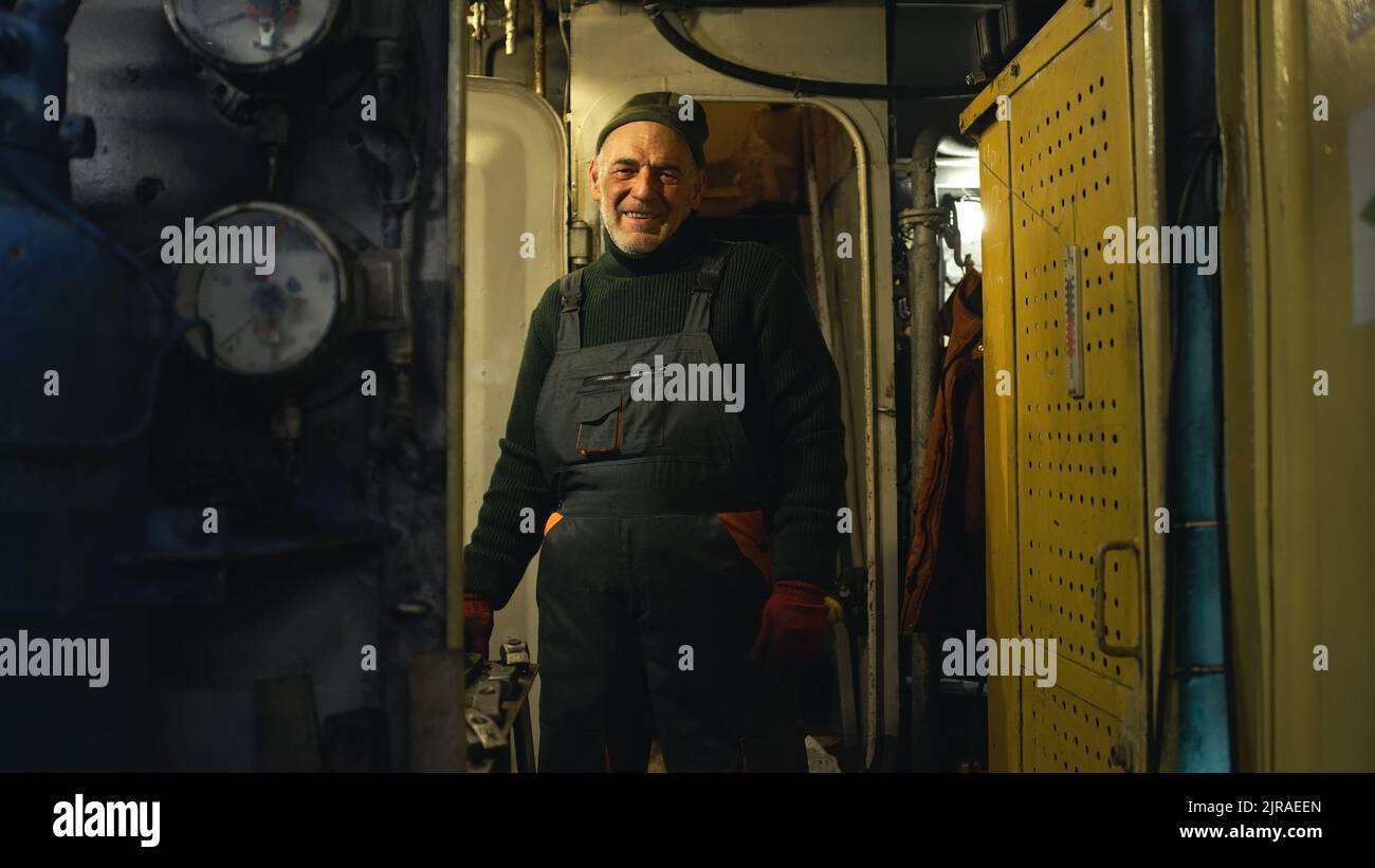 Zoom out-Ansicht eines älteren Mannes, der lächelt und anfängt, den Hebel zu drehen, während er hinter der Tür des Maschinenraums des Fischerschiffes steht Stockfoto