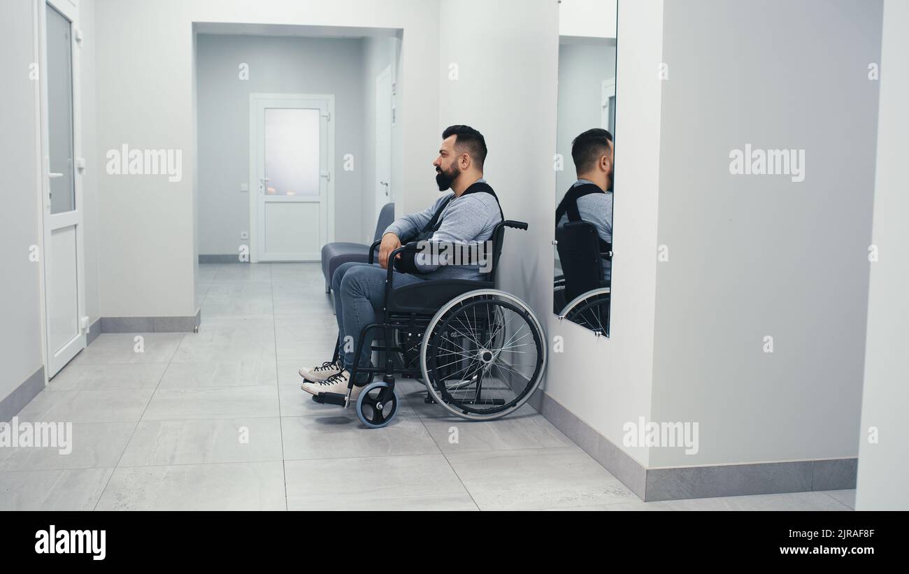 Behinderter Mann mit gebrochenem Arm sitzt auf dem Rollstuhl und wartet auf dem Flur des modernen Krankenhauses Stockfoto