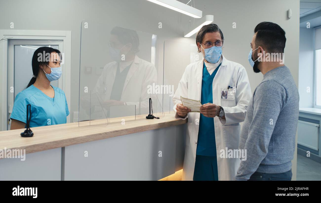 Asiatische Frau in Maske im Gespräch mit Mann dann Papier an herannahenden reifen Arzt während der Arbeit an der Aufnahme in modernen Krankenhaus während Pandemie Stockfoto