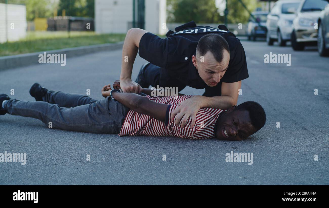 Aggressive Frau in Polizeiuniform, die schwarzen Mann zu Boden drückt und während der Verhaftung auf der Straße redet Stockfoto