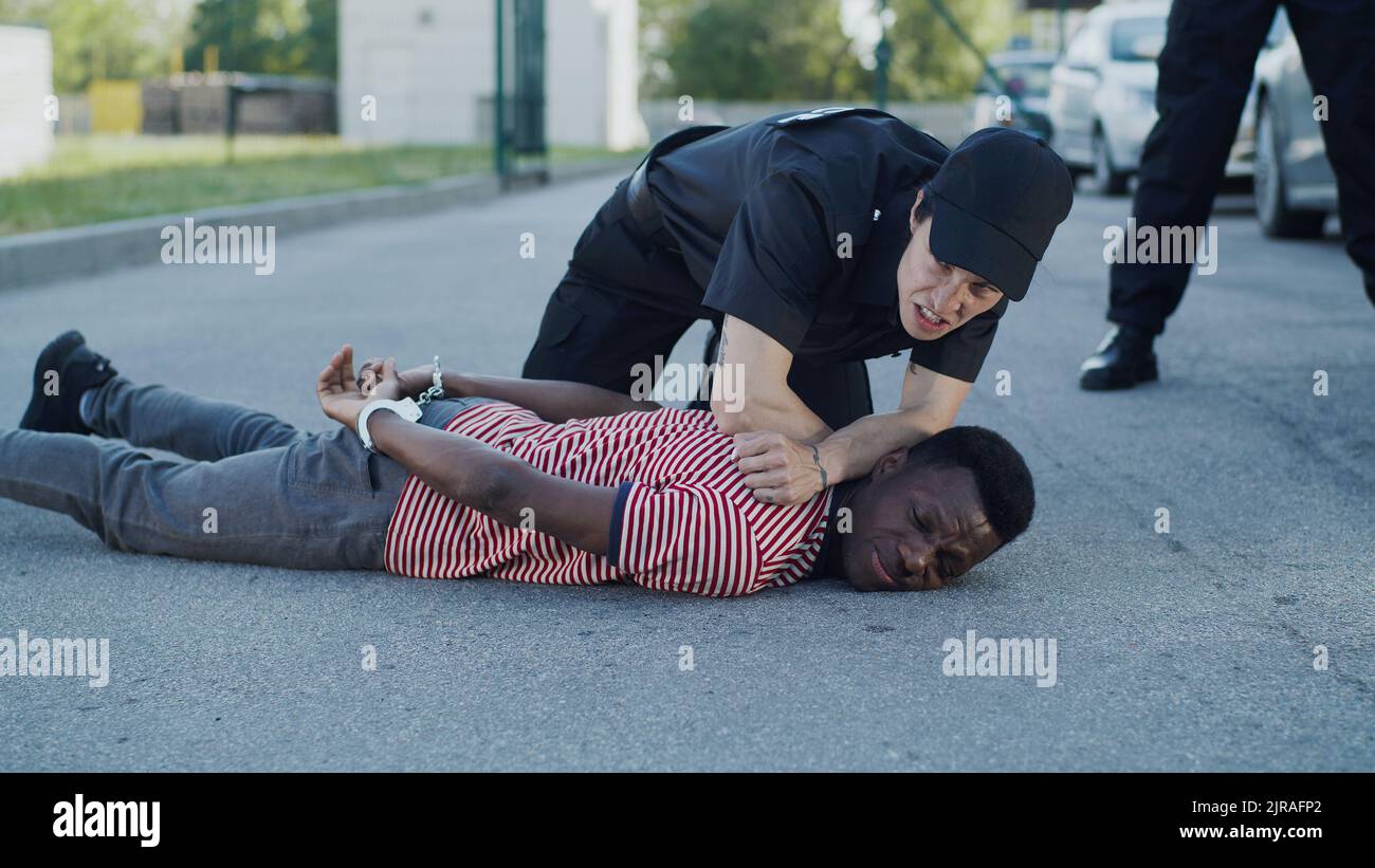 Afroamerikanischer Polizist, der während der Festnahme hinter Gitter mit schwarzer Sträfin in der Nähe einer Kollegin mit Pistole spricht Stockfoto