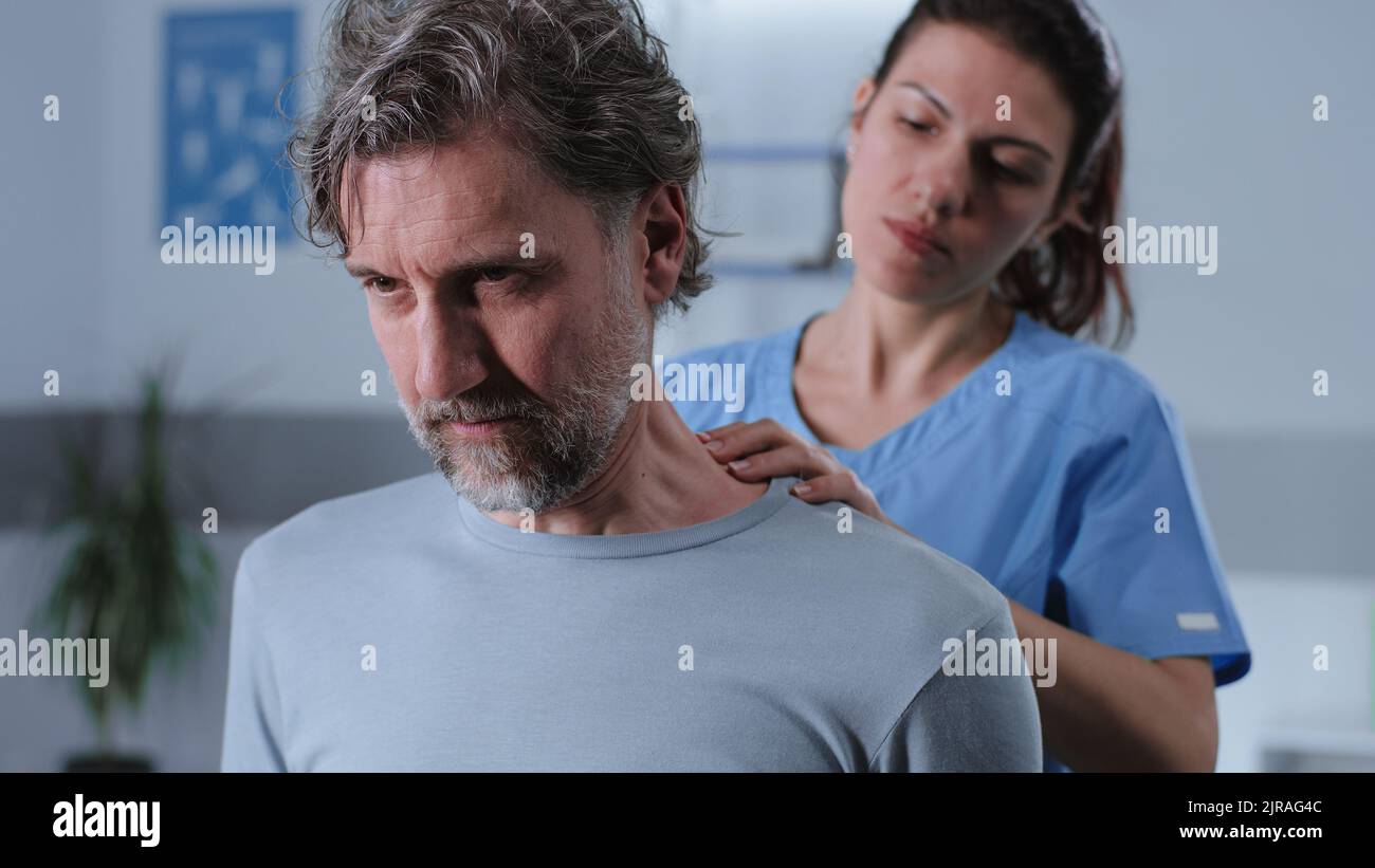 Erwachsene Frau in medizinischer Uniform massiert Hals und Schultern von bärtigen männlichen Patienten mittleren Alters während der Arbeit in der Rehabilitationsklinik Stockfoto