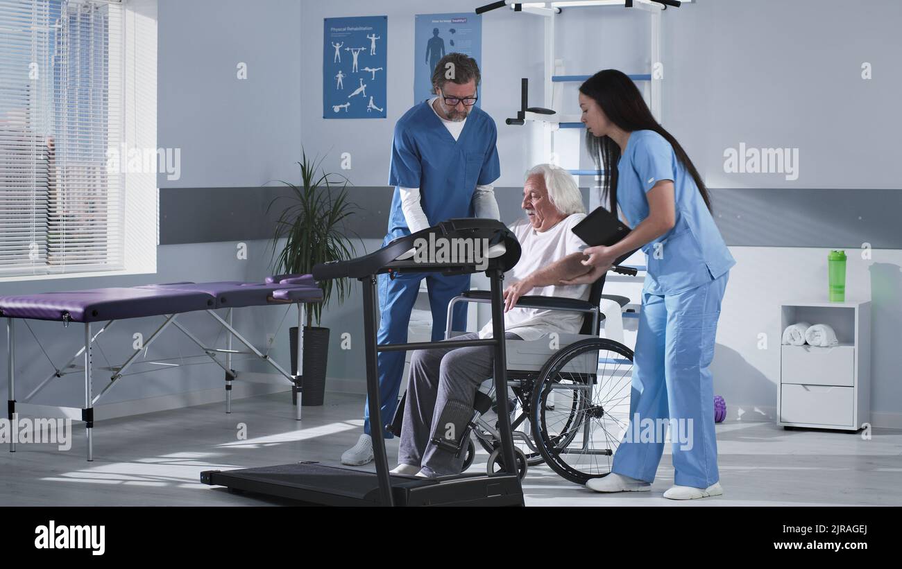 Ärzte helfen älteren Menschen, auf dem Laufband zu klettern und erklären Rehabilitationsübungen während der Arbeit in einer modernen Klinik Stockfoto