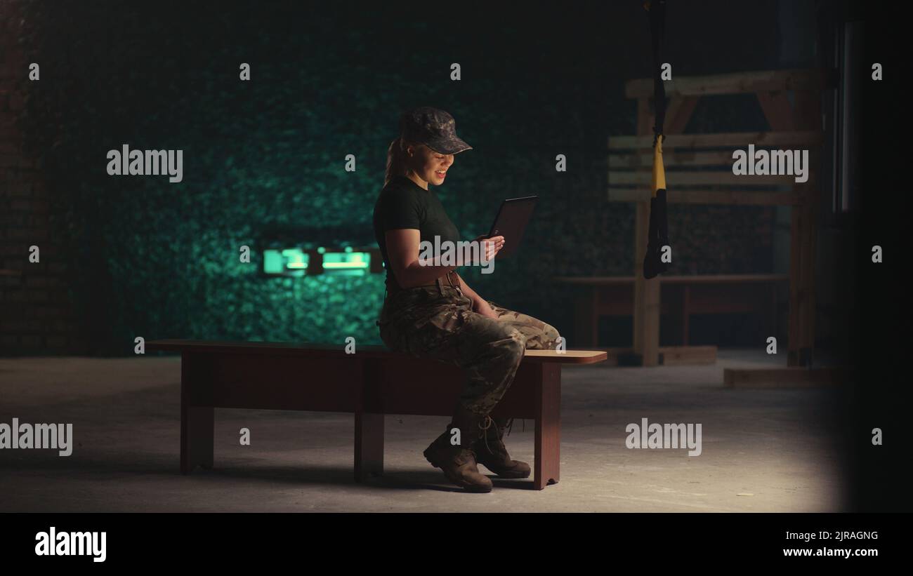 Blick auf die entzückte Soldatin, die lächelt und mit der Hand winkt, während sie auf der Bank sitzt und auf dem Tablet in der dunklen Turnhalle des Militärstützpunktes einen Videoanruf abruft Stockfoto