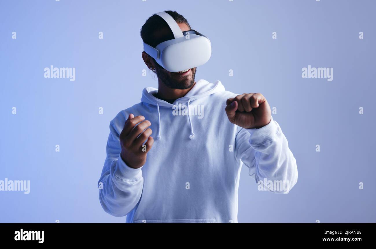Fröhlicher junger Mann, der fröhlich lächelt und dabei ein immersives Virtual Reality-Erlebnis hat. Junger Mann, der durch virtuelle Köpfe auf seine Faust blickt Stockfoto
