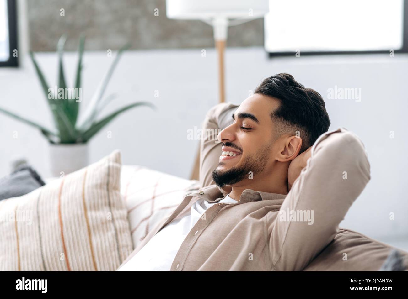 Nahaufnahme eines glücklichen, ruhigen indischen oder arabischen jungen Mannes, der sich im Wohnzimmer auf dem Sofa ausruht, seine Hände mit geschlossenen Augen hinter seinen Kopf hält und lächelt. Entspannen und entspannen zu Hause. Stockfoto