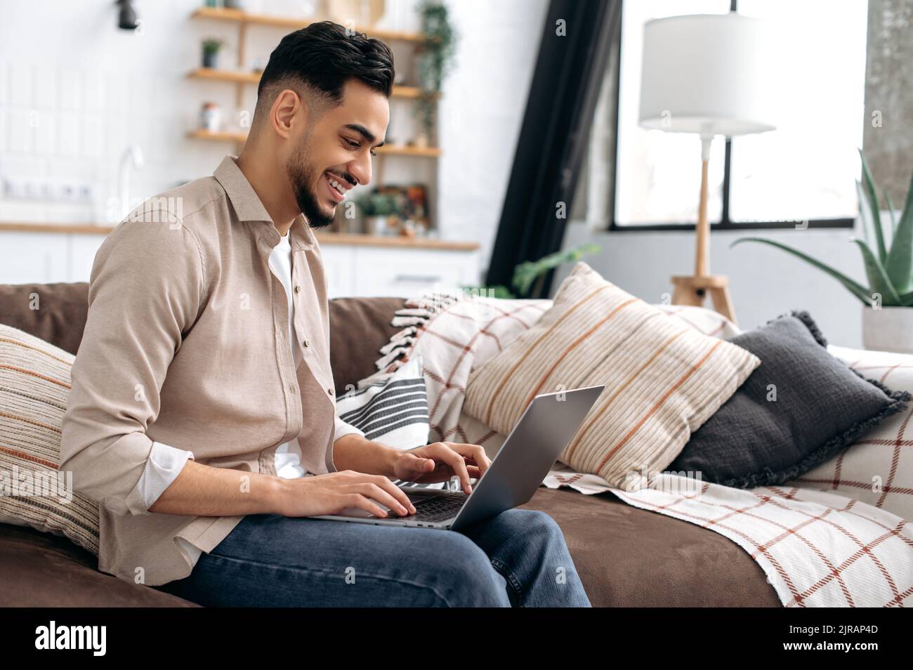 Frohes, positives arabisches oder indisches, junger, moderner Mann, der Zeit zu Hause verbringt, auf dem Sofa im Wohnzimmer sitzt, Laptop benutzt, im Internet surft, soziale Medien anschaut, Videos anschaut, Lächelnd Stockfoto