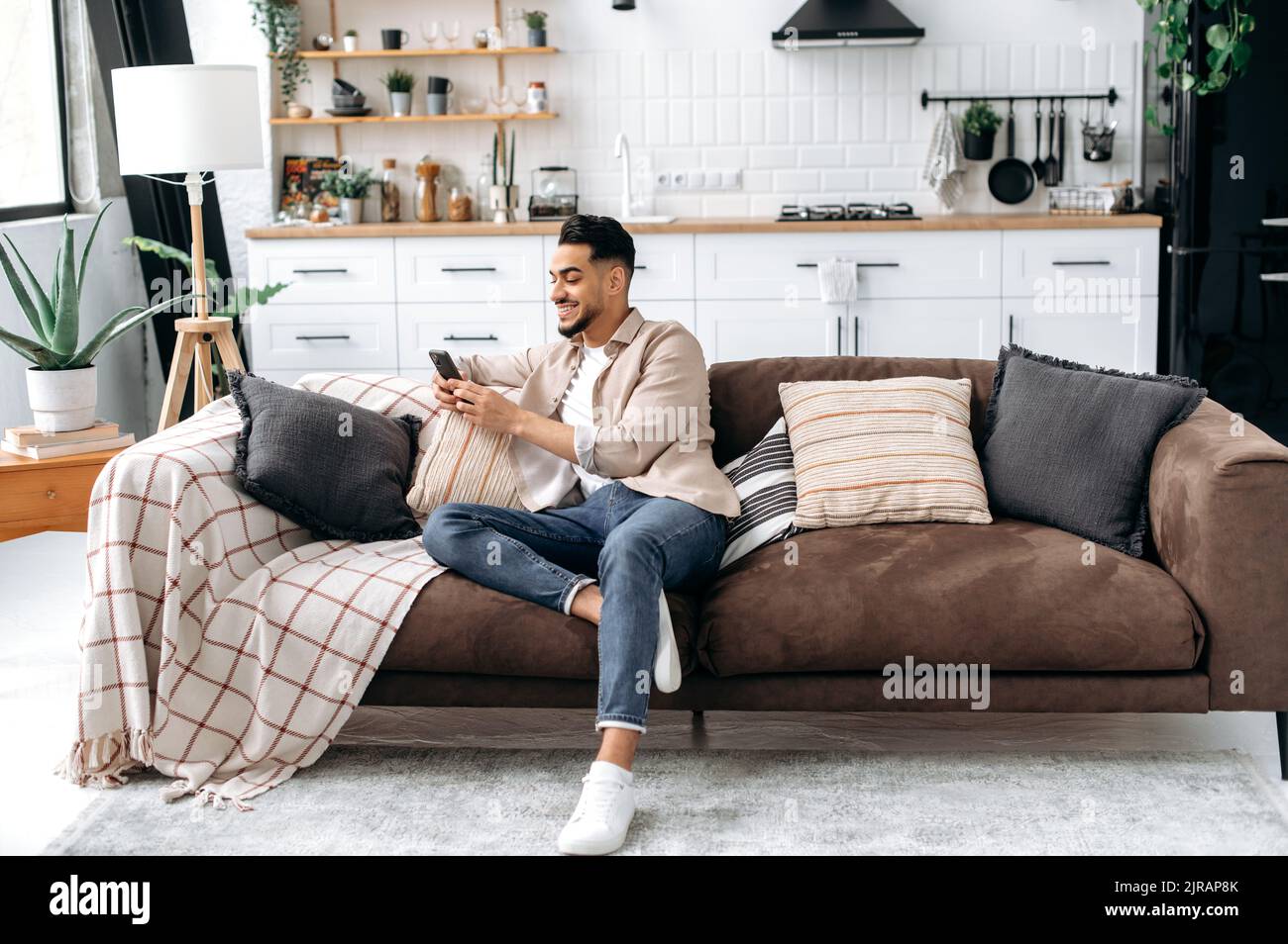 Online-Messaging, Wireless-Technologie. Fröhlicher inder oder araber, sitzen im modernen Wohnzimmer auf einem bequemen Sofa, benutzen sein Smartphone, surfen und Messaging in einem Social Media, surfen im Internet, lächeln Stockfoto