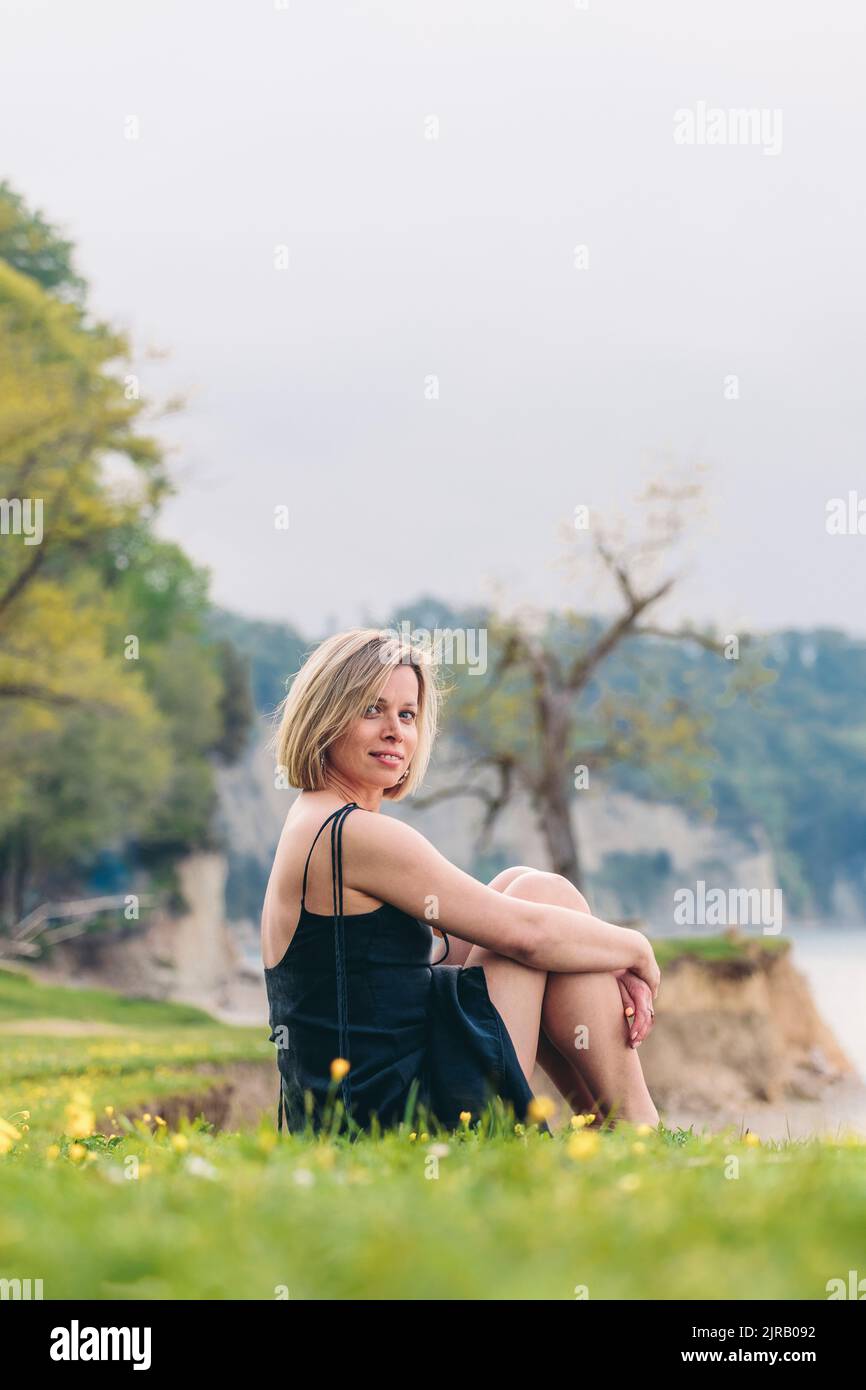 Lächelnde reife Frau mit blonden Haaren auf Gras sitzend Stockfoto