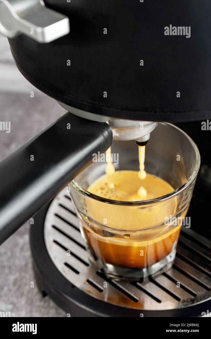 Glasbecher steht auf dem Gitter der schwarzen Kaffeemaschine und gießt Kaffee hinein. Prozess der Zubereitung von Kaffee. Stockfoto