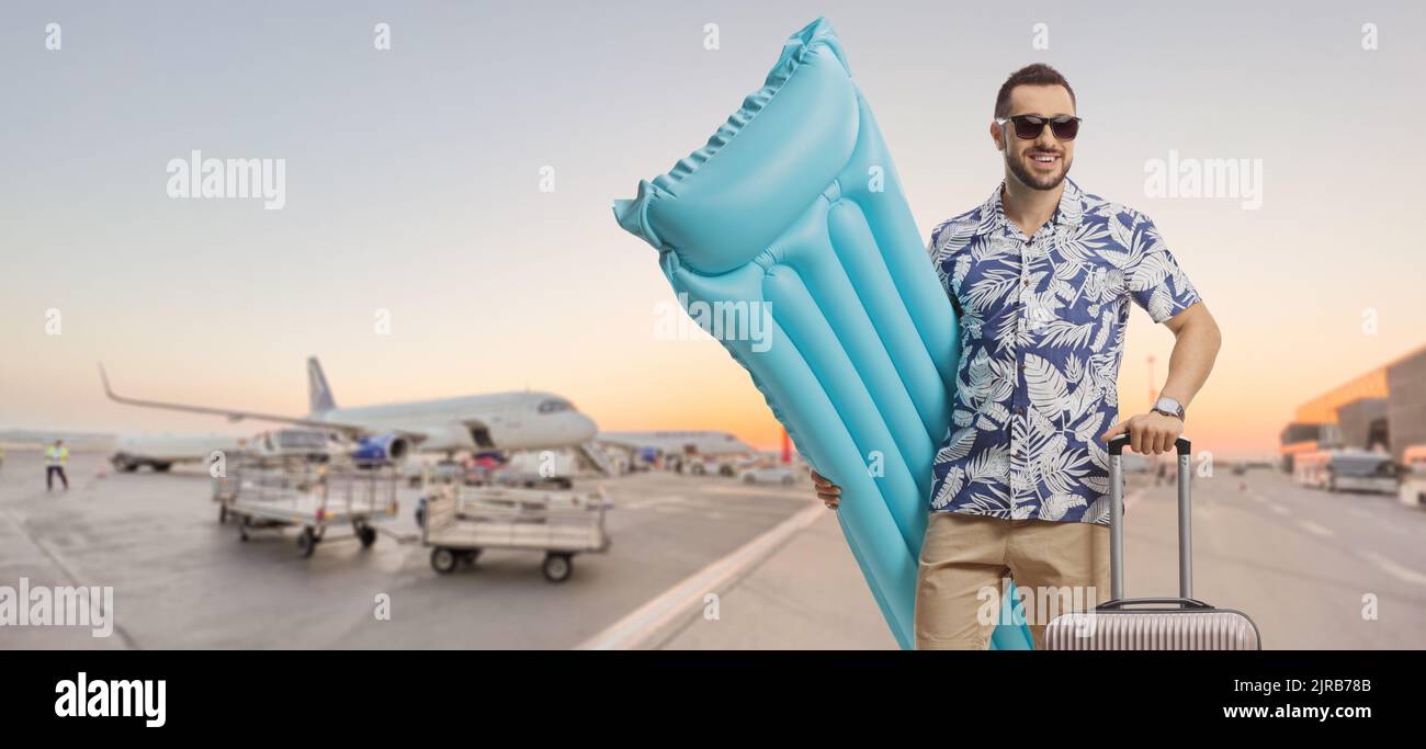 Junger männlicher Tourist, der eine aufblasbare Matratze hält und auf einer Start- und Landebahn des Flughafens posiert Stockfoto