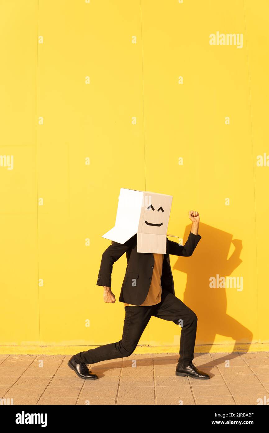 Mann trägt eine Schachtel mit einem Gesicht, das vor der gelben Wand auf dem Fußweg sticht Stockfoto