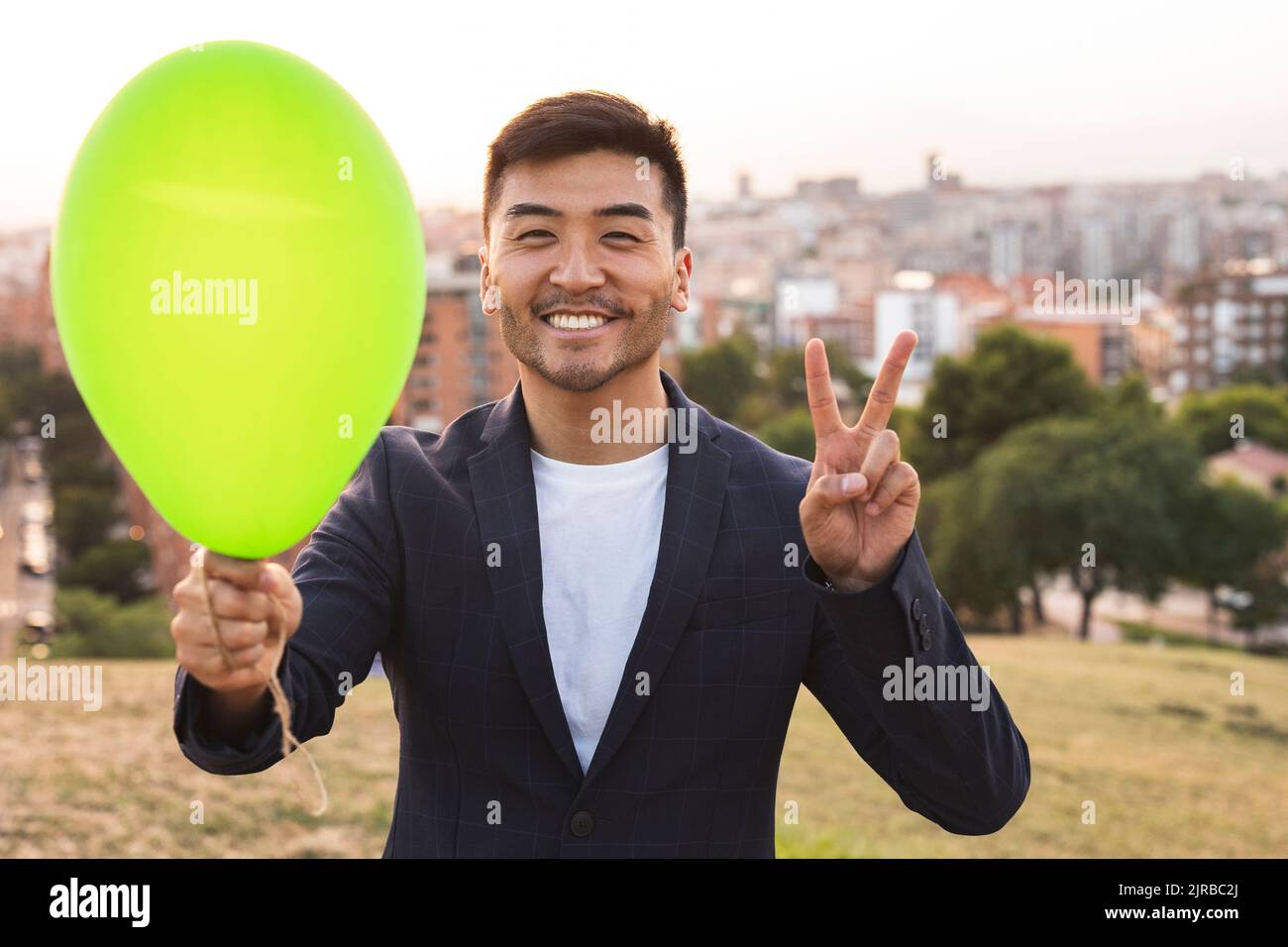Fröhlicher junger Geschäftsmann mit grünem Ballon, der bei Sonnenuntergang ein Friedenszeichen zeigt Stockfoto