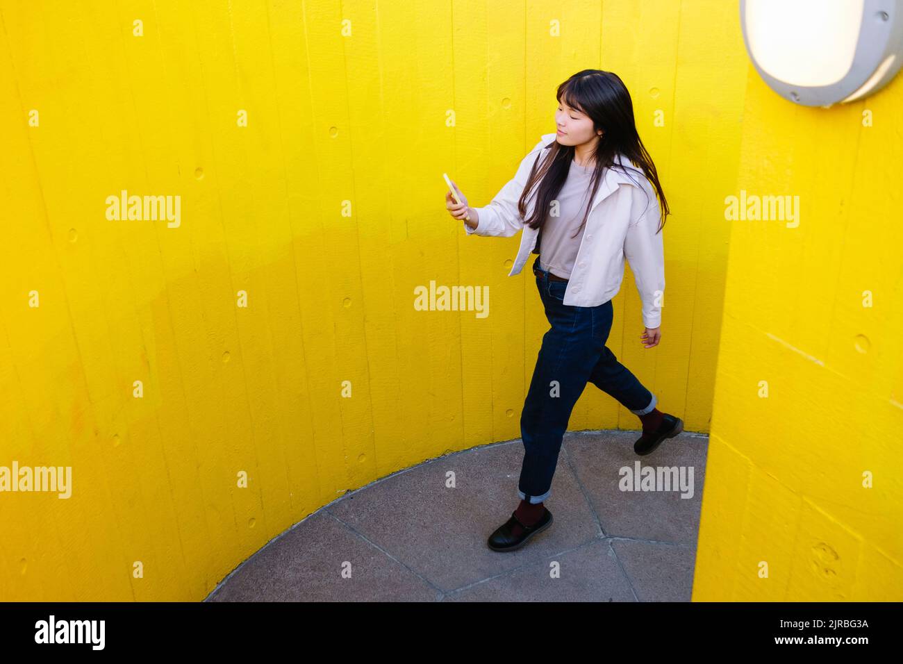 Junge Frau, die mit dem Mobiltelefon unterwegs ist, läuft zwischen gelben Wänden Stockfoto