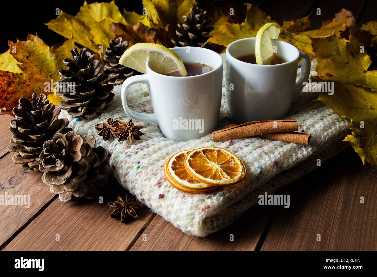 Zwei Tassen Tee mit Zitrone auf einem warmen gestrickten Winterschal. Herbstliche Komposition auf einem Holztisch, mit Zimtstangen, Sternanise, Zapfen und Blättern. Stockfoto