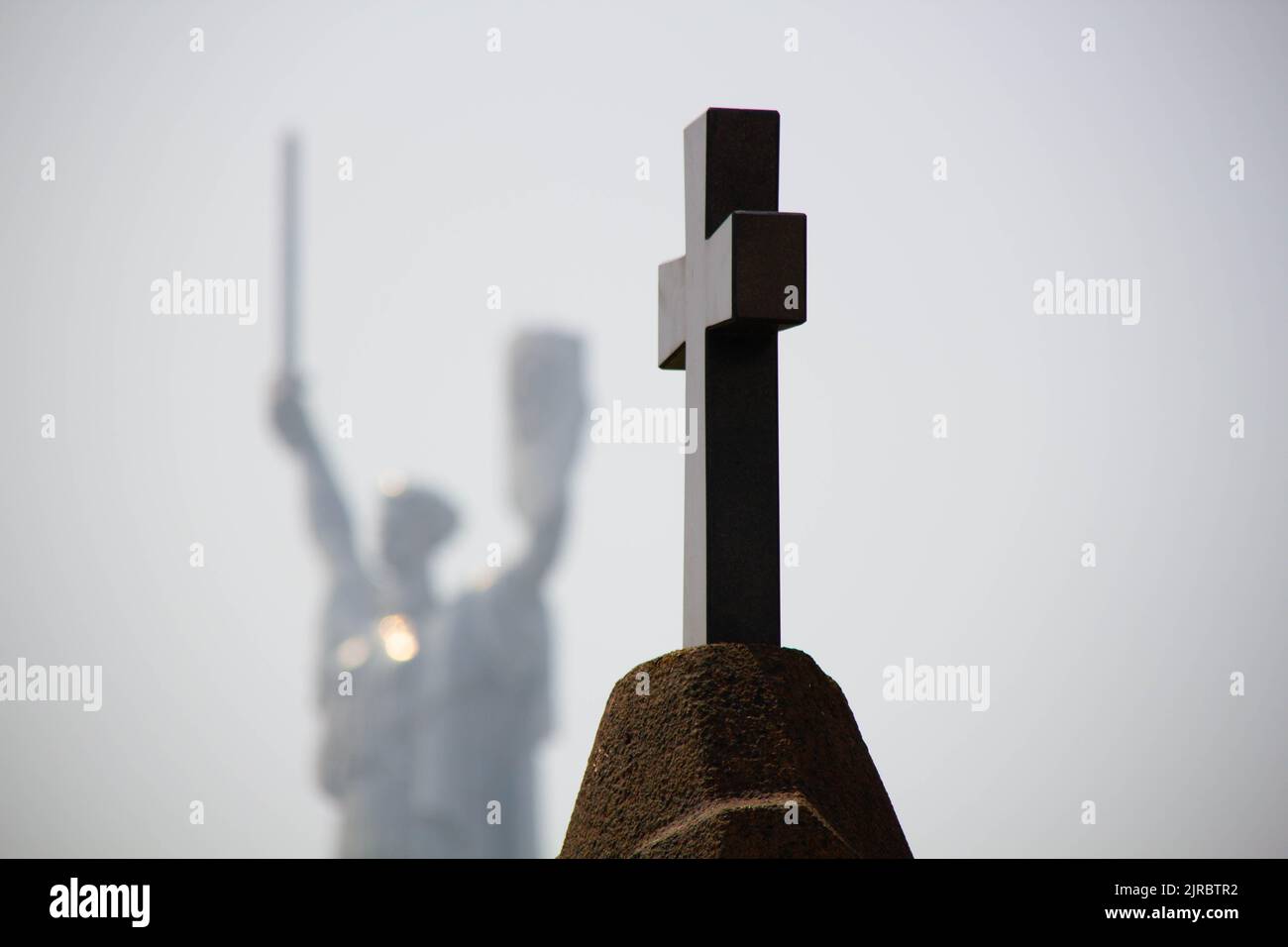 Überqueren Sie den Friedhof. Ein katholisches Kreuz auf dem Grab vor dem Hintergrund eines Militärdenkmals für das Mutterland. Christliches Grabkreuz Stockfoto