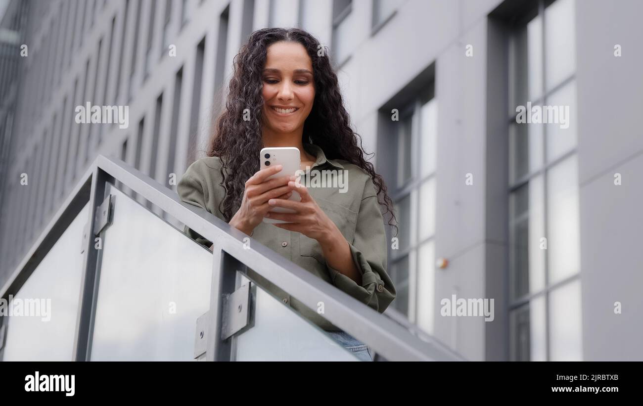 Lächelnd kaukasisch hispanisch glücklich zufrieden Frau Benutzer Dame stehen un Stadt im Freien auf Treppen chatten SMS suchen im Netz mit Handy App Telefon Stockfoto