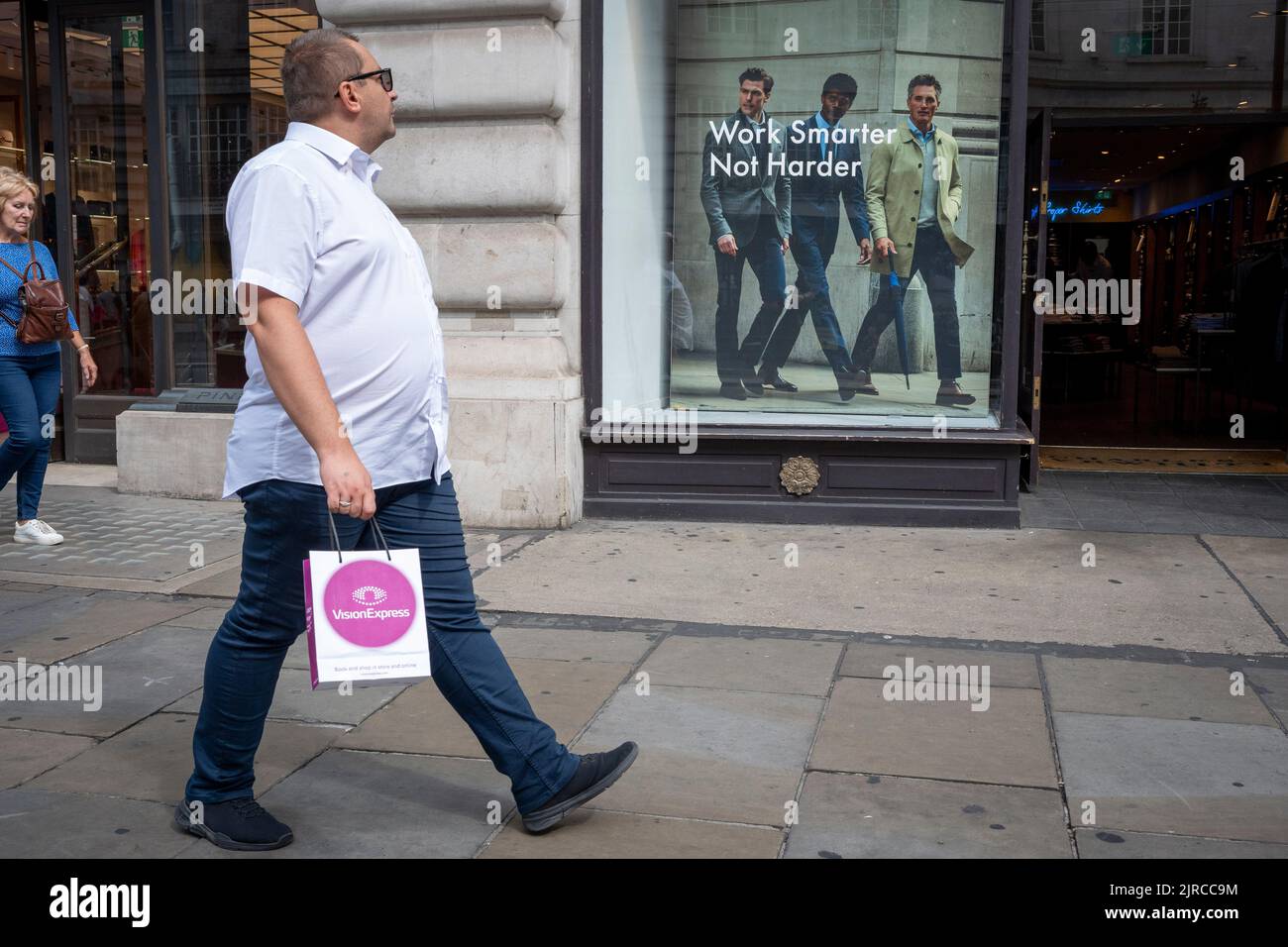 Die Käufer gehen am Fenster des Herrenmode-Einzelhändlers Charles Tyrwhitt vorbei, dessen arbeitsethischer Slogan britischen Arbeitnehmern sagt, dass sie „intelligenter arbeiten sollten, nicht härter“ - eine Idee, die der Favoritin der Kandidaten der konservativen Führung, Liz Truss, widerhallt, die am 23.. August 2022 behauptet hat, dass die britische Belegschaft faul sei, In London, England. In einem durchgesickerten Gespräch sagt Truss, dass britische Arbeiter „mehr Transplantat“ brauchen. Stockfoto