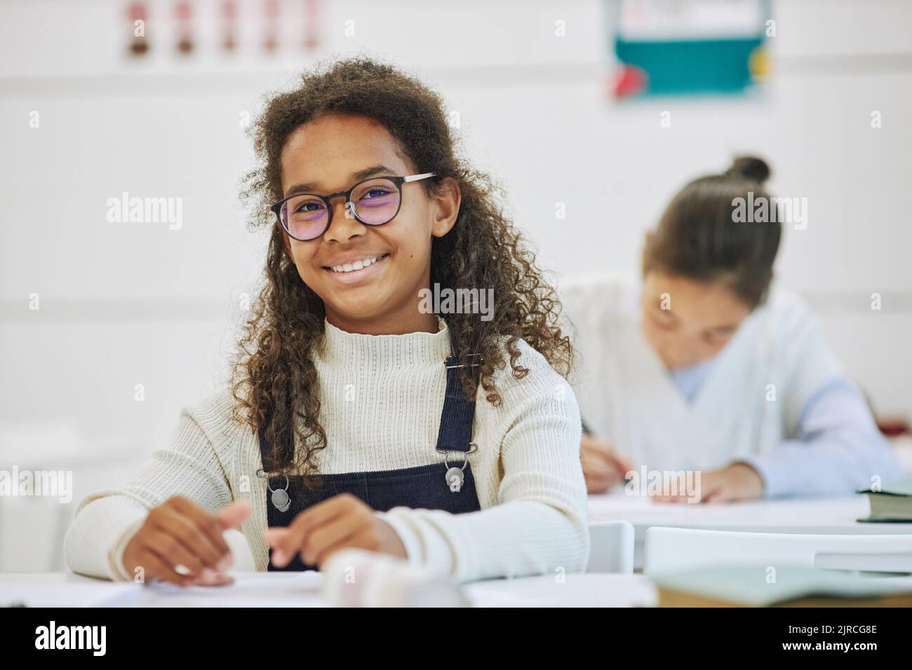 Porträt eines lächelnden schwarzen Schulmädchen mit Brille und Blick auf die Kamera, während sie am Schreibtisch in der Schule sitzt, Platz kopieren Stockfoto