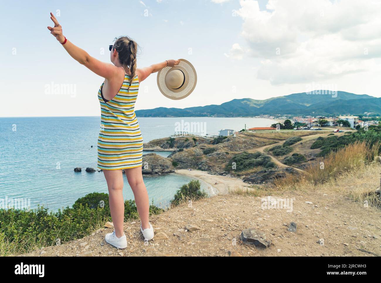 Junge, fröhliche kaukasische Frau in einem gestreiften Kleid, die ihre Arme streckt, während sie die wunderschöne griechische Meereslandschaft bewundert. Urlaubskonzept. Hochwertige Fotos Stockfoto