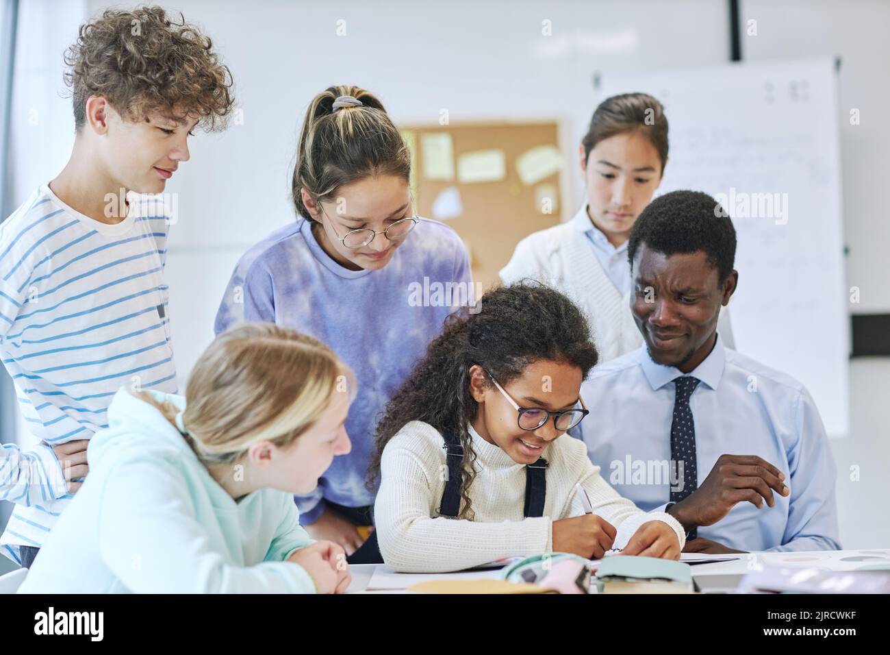 Eine vielfältige Gruppe von Kindern, die in der Schule fröhlich lächeln, während sie mit einem jungen Lehrer zusammenarbeiten Stockfoto