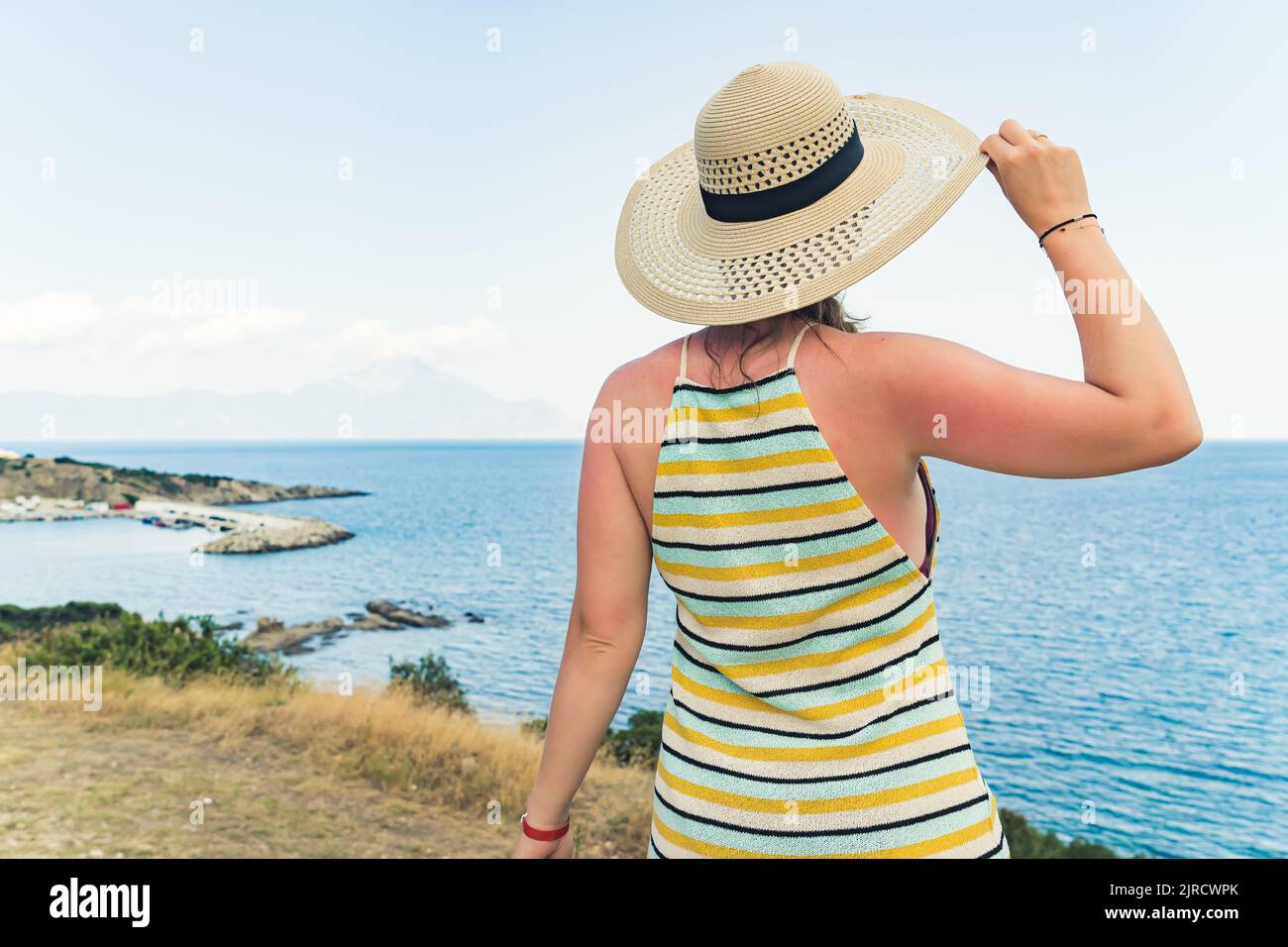 Wunderschöne, klare Küste, die von einem kaukasischen Touristen in einem ärmellosen gestreiften Kleid und einem beigen Sommerhut bewundert wird, der rückwärts vor die Kamera steht. Tagsüber. Blauer Himmel. Hochwertige Fotos Stockfoto