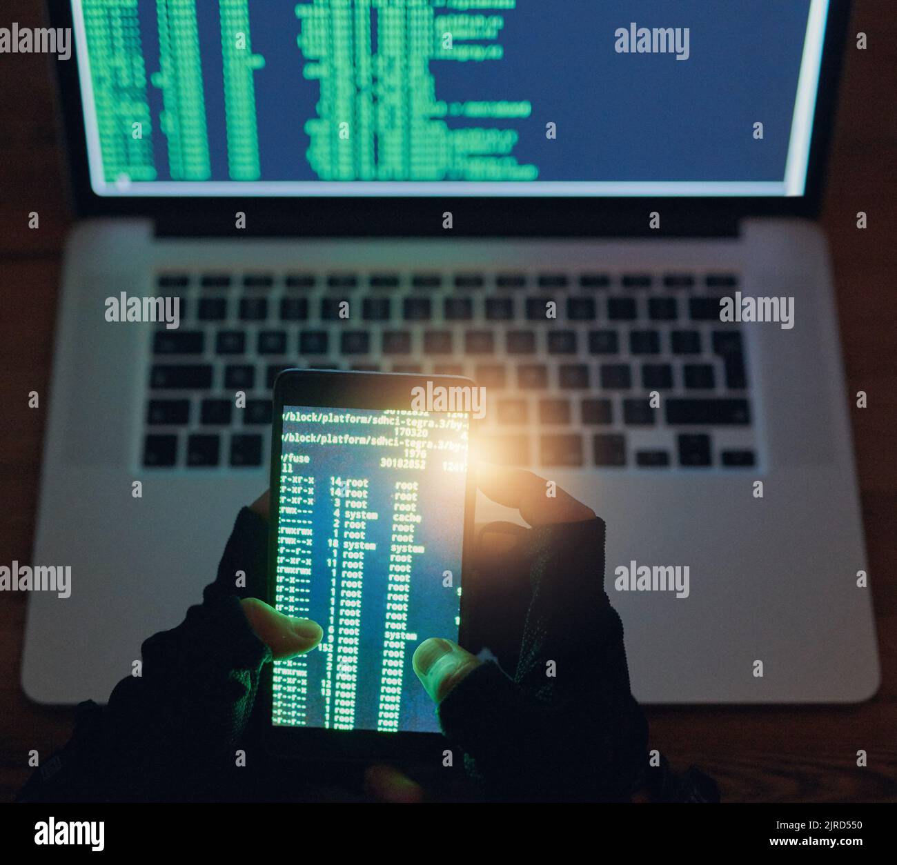 Durchbrechen mehrerer Netzwerke. Ein nicht erkennbarer Hacker, der im Dunkeln ein Handy und einen Laptop benutzt. Stockfoto