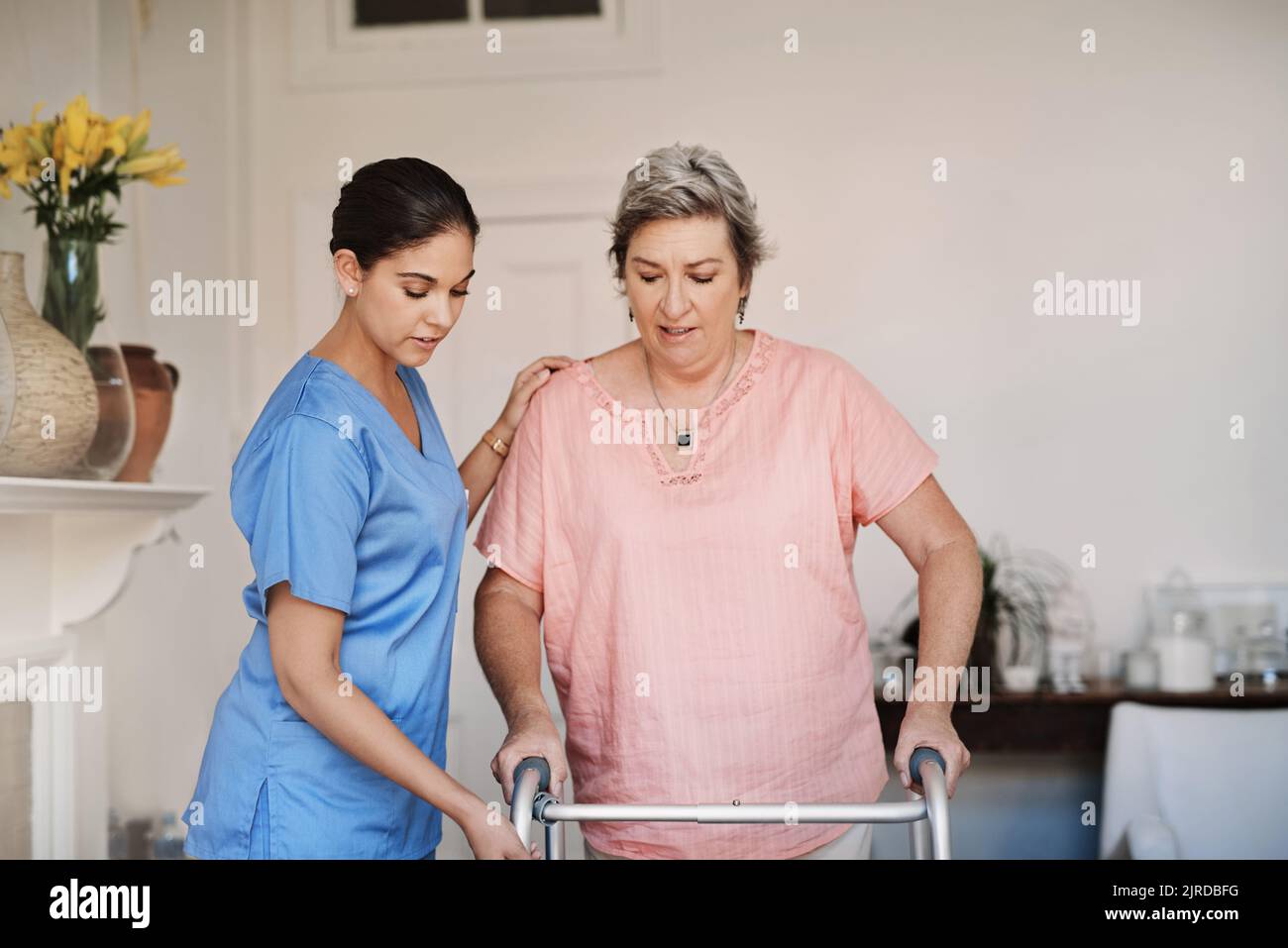 Langsam und stetig Schritte jetzt ... eine attraktive junge Betreuerin, die einem älteren Patienten mit einem Geher in einem Pflegeheim hilft. Stockfoto