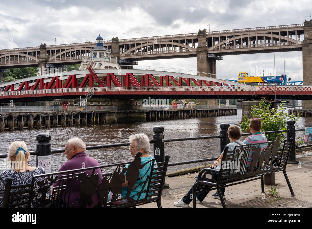 Menschen sitzen auf Bänken am Kai, Newcastle upon Tyne, Großbritannien, neben der Swing Bridge mit dem Toon Tour Bus, der quer fährt. Stockfoto
