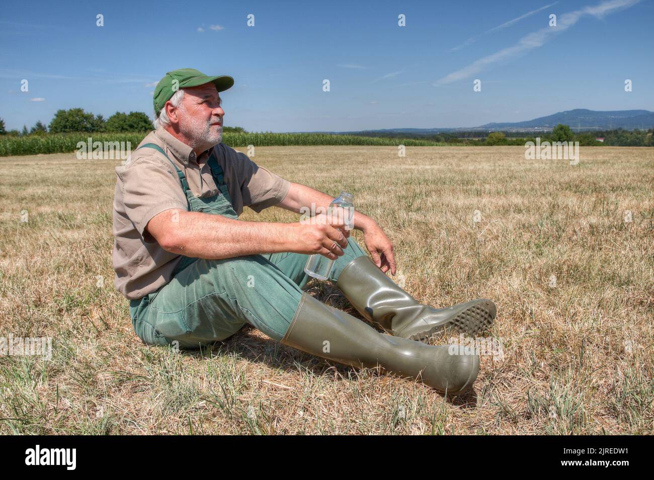 Ein Bauer sitzt hilflos auf seiner ausgetrocknten Wiese mit einer Flasche Wasser in der Hand und blickt nachdenklich in den Himmel. Stockfoto