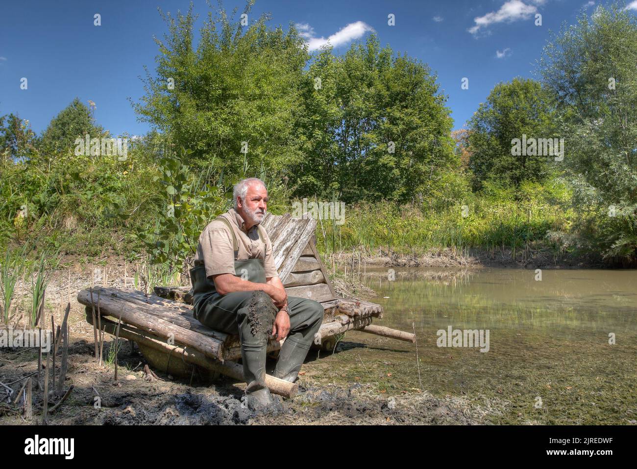 Ein Teichwirt sitzt auf trockenem Land und schaut ängstlich auf den Rest des Wassers. Die anhaltende Hitze lässt das kleine Biotop austrocknen. Stockfoto