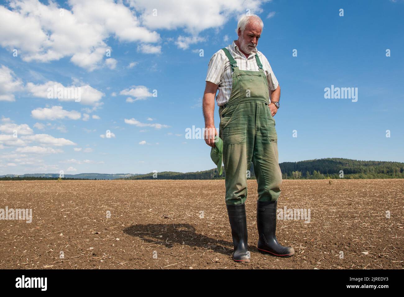Ein erfahrener Bauer in grünen Latzhose und schwarzen Gummistiefeln steht auf dem frisch gesät Feld und schaute ängstlich auf die trockene Erde. Stockfoto