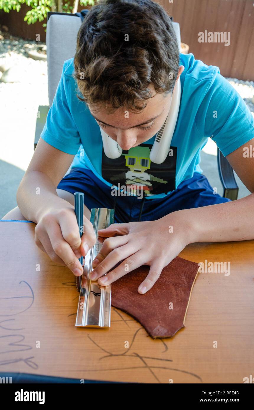 Ein Teenager verwendet ein scharfes Skalpell und ein Metalllineal als Anleitung, um ein gerades Stück Leder zu schneiden. Stockfoto